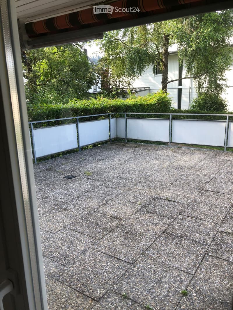 Empty balcony with a concrete floor and white privacy screens, overlooking a garden area with trees and bushes.