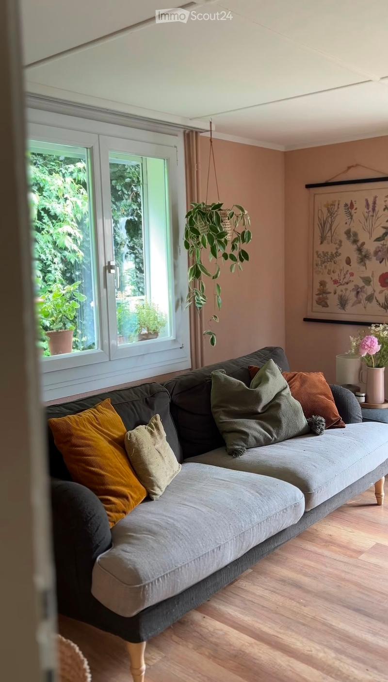 A cozy living room with a grey sectional sofa adorned with multiple pillows, a hanging plant, a potted plant on a side table, and a vase with flowers on a small table. The room features light wooden flooring and large windows with a view of greenery outsi