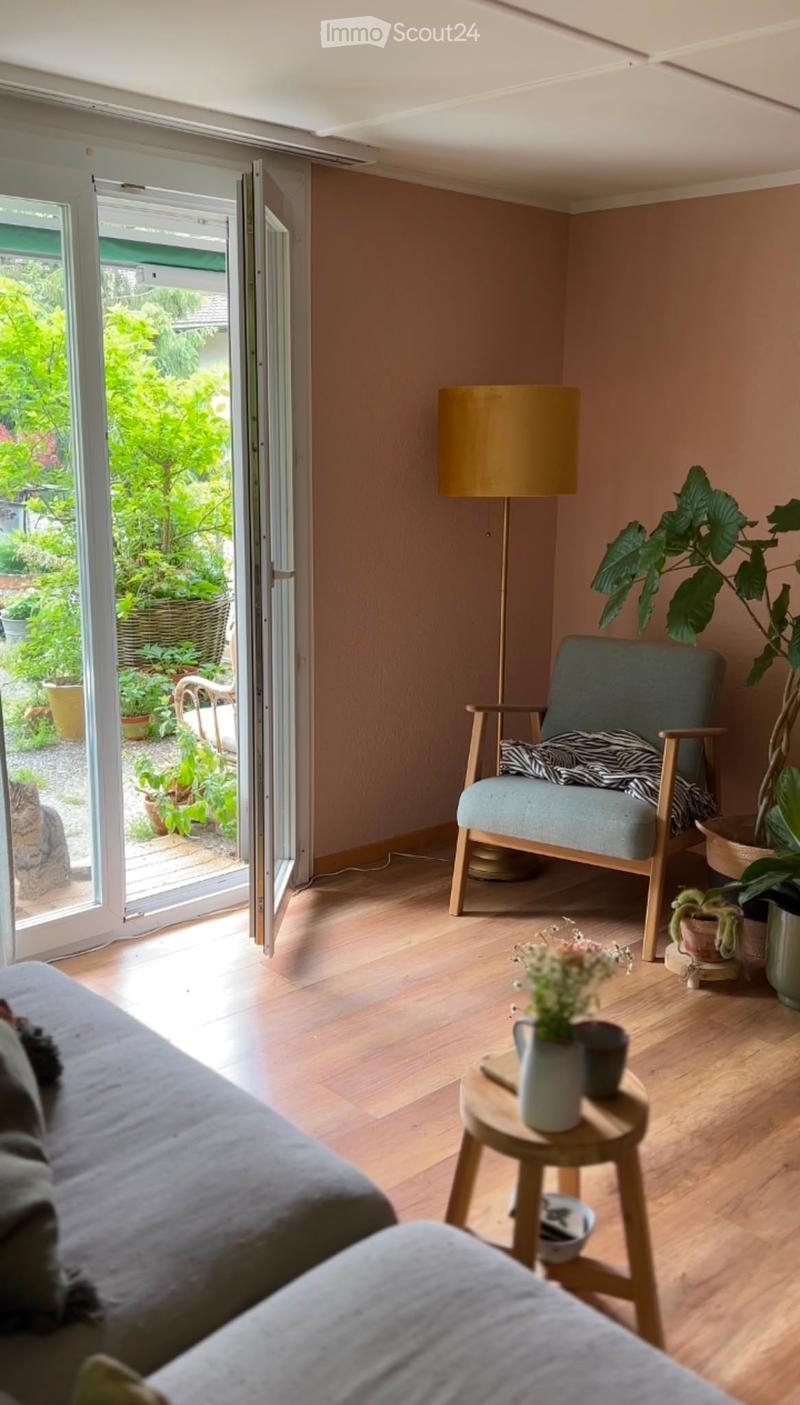 Living room with wooden flooring, a grey sofa, a wooden chair with a cushion, a small round stool with a flower pot, and a floor lamp. A cat is visible near the open sliding doors leading to a garden.