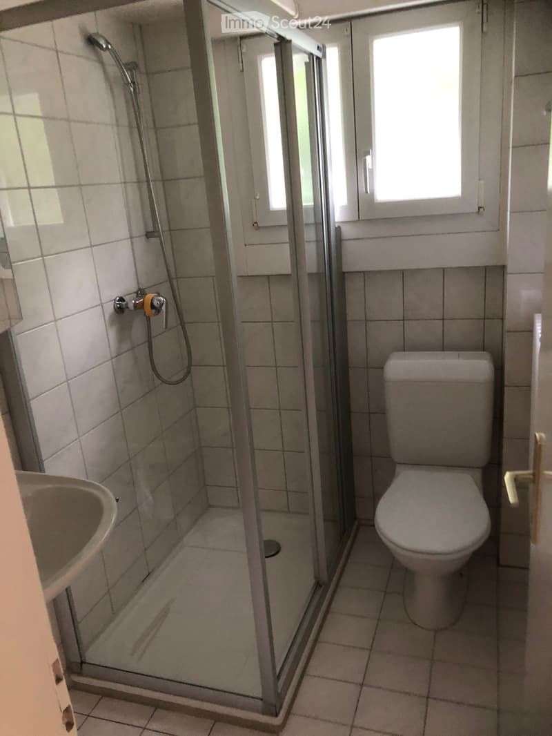 Bathroom with a toilet, sink, and a glass-enclosed shower area. The walls and floor are tiled in light gray.