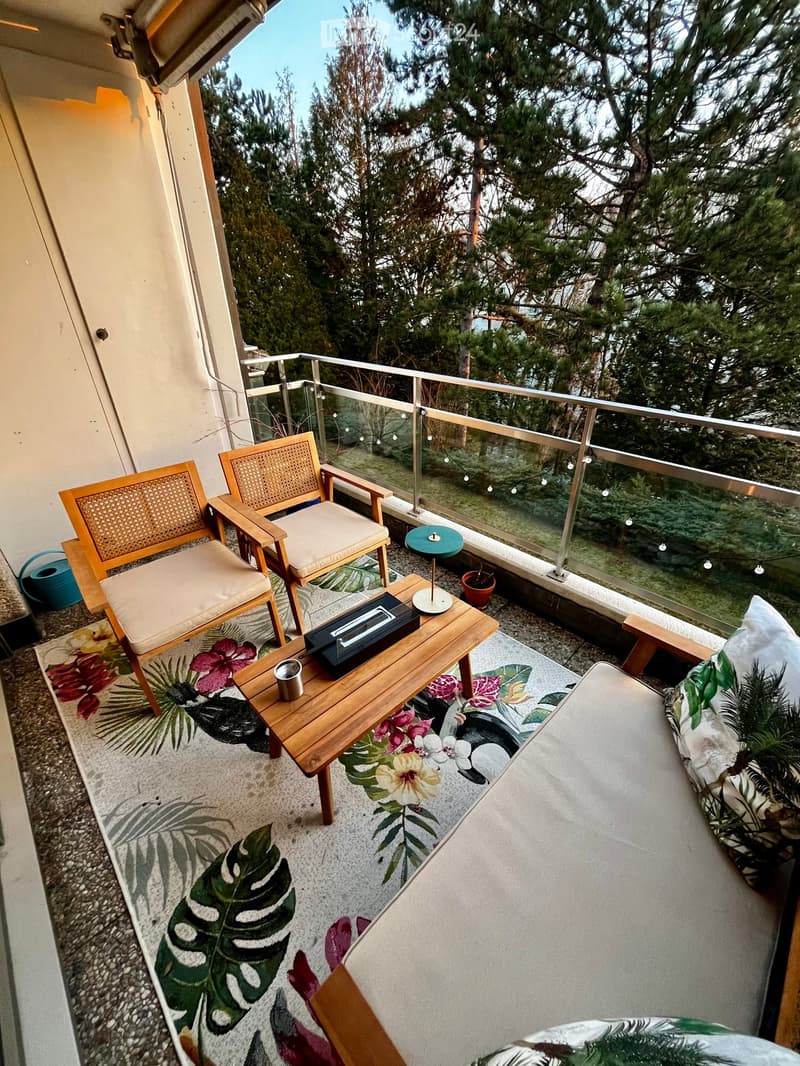 Balcony / Terrace. Balcony with wooden chairs, a small table, a sofa, and a rug, overlooking a green, tree-filled area.