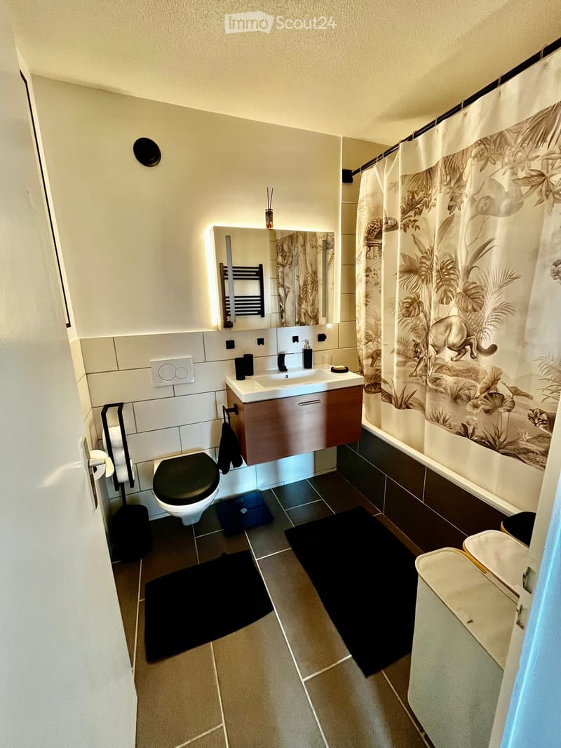 Bathroom. Modern bathroom with a toilet, sink, mirror, and shower curtain. Features a wooden vanity cabinet, black tiles, and a decorative mirror with LED lighting.