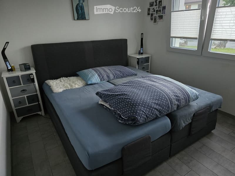 Modern bedroom with a large bed, nightstands, and decorative pillows. The room features a large window with blinds and a cozy, contemporary design.
