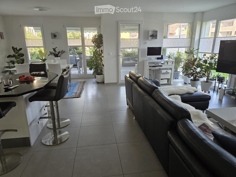 Modern living room with a black leather sofa, a white fur throw, a glass table, and several bar stools. Large windows and sliding glass doors lead to a balcony area with potted plants.