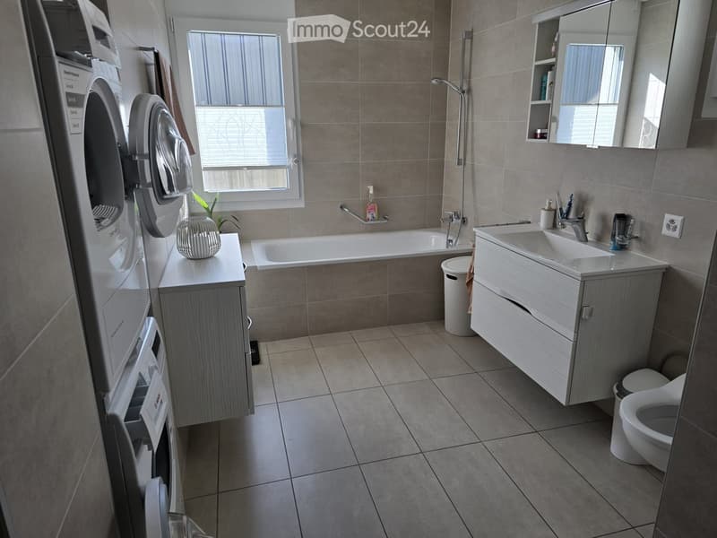 Modern bathroom with beige tiles, a bathtub, a sink with a cabinet, a toilet, a washing machine, a mirror, and a small plant on a cabinet.