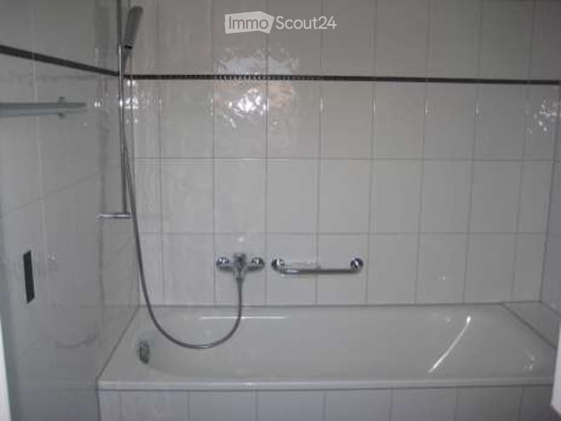 Bathtub with a handrail, shower hose, and faucet in a tiled bathroom.