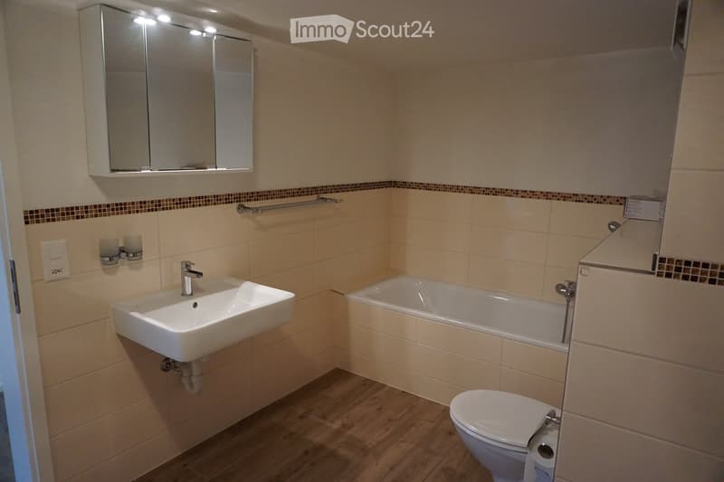 Modern bathroom with beige tiles, a white sink, a bathtub, a toilet, a mirror cabinet, and a towel rack.