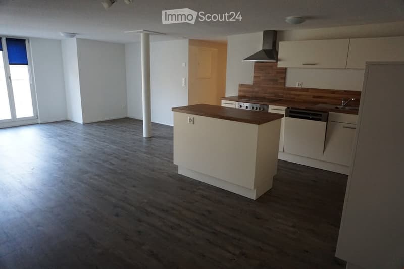 Modern kitchen with light-colored cabinets, a central island, and built-in appliances including a stove, dishwasher, and sink. The space features wooden flooring and ample natural light from a large window.
