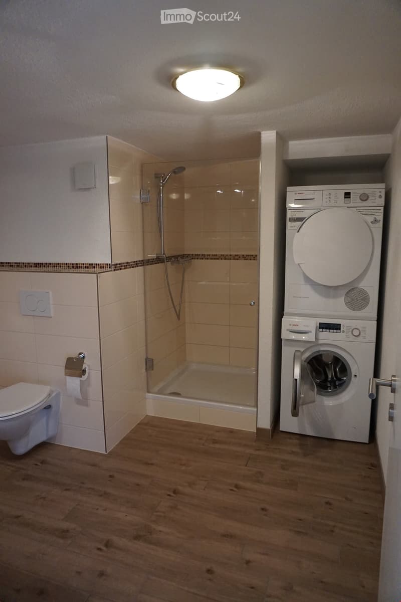 Modern bathroom with a toilet, glass-enclosed shower, and stacked Bosch washing machine and dryer unit. Light wood flooring and beige tiles.