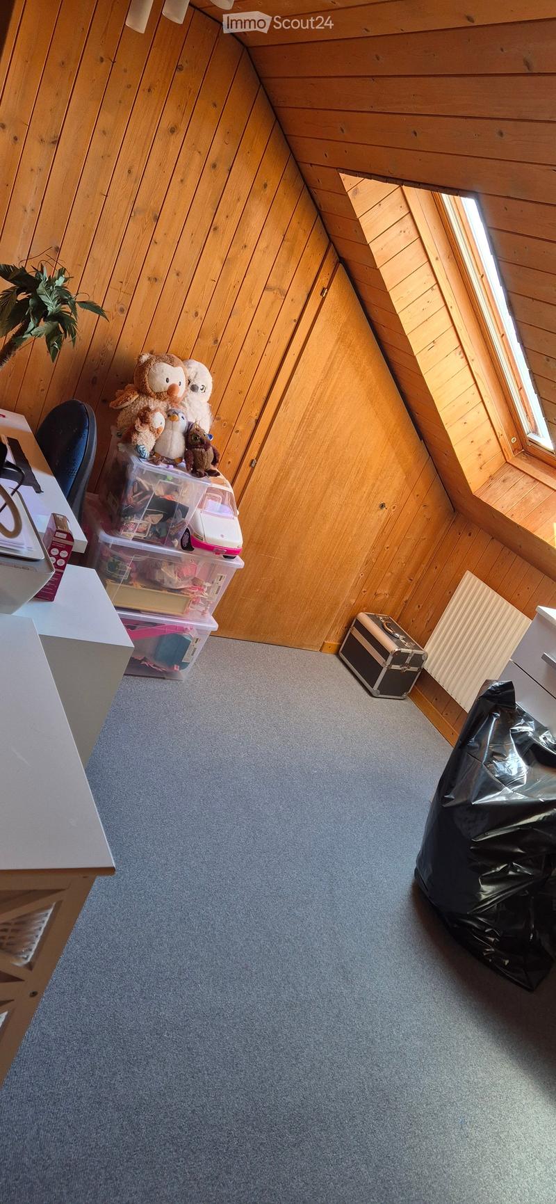 A cozy attic room with wooden walls and a slanted ceiling, featuring a grey carpeted floor, a white desk with a chair, storage bins filled with toys and stuffed animals, a small suitcase, and a black trash bag.