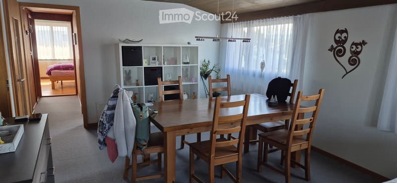 Bright dining area with a wooden dining table and chairs, a white shelving unit with decorative items, and a view into a bedroom with a bed and pink bedding.