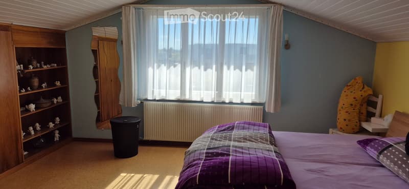 Cozy bedroom with a bed, purple bedding, a wooden shelf with decorative items, a mirror, a radiator, and a yellow bean bag chair.