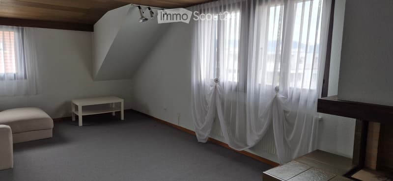 Bright, carpeted attic room with white walls, large windows with sheer curtains, a small white table, and a beige sofa.