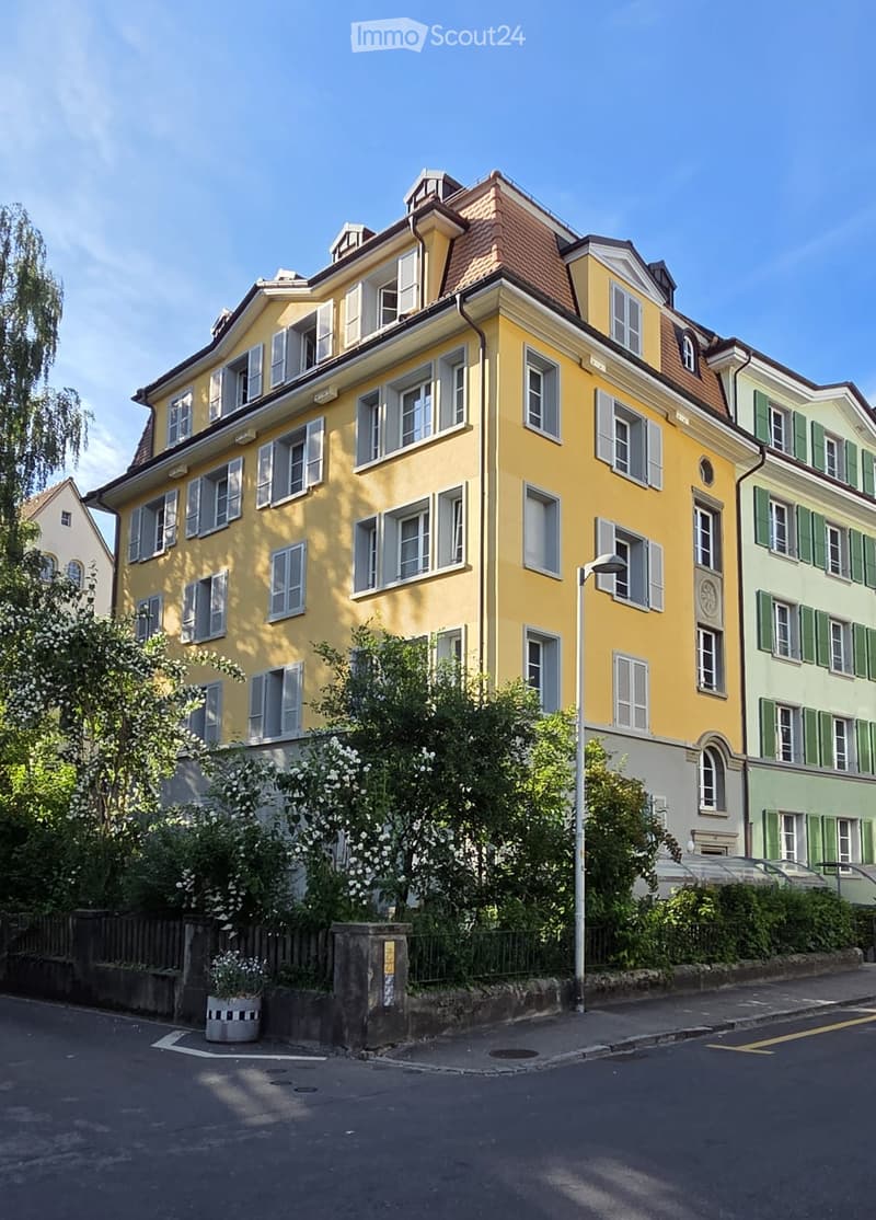 A multi-story yellow building with white shutters and a red-tiled roof, surrounded by greenery and neighboring houses.