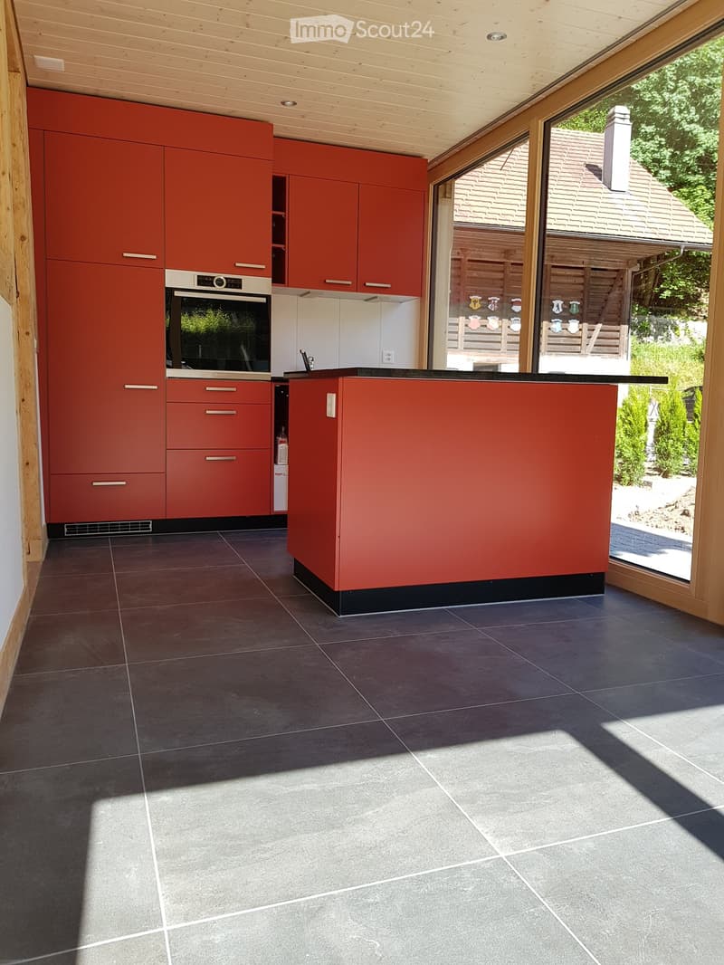 Modern kitchen with red cabinets, a built-in oven, and a central island, featuring large windows and a wooden ceiling.