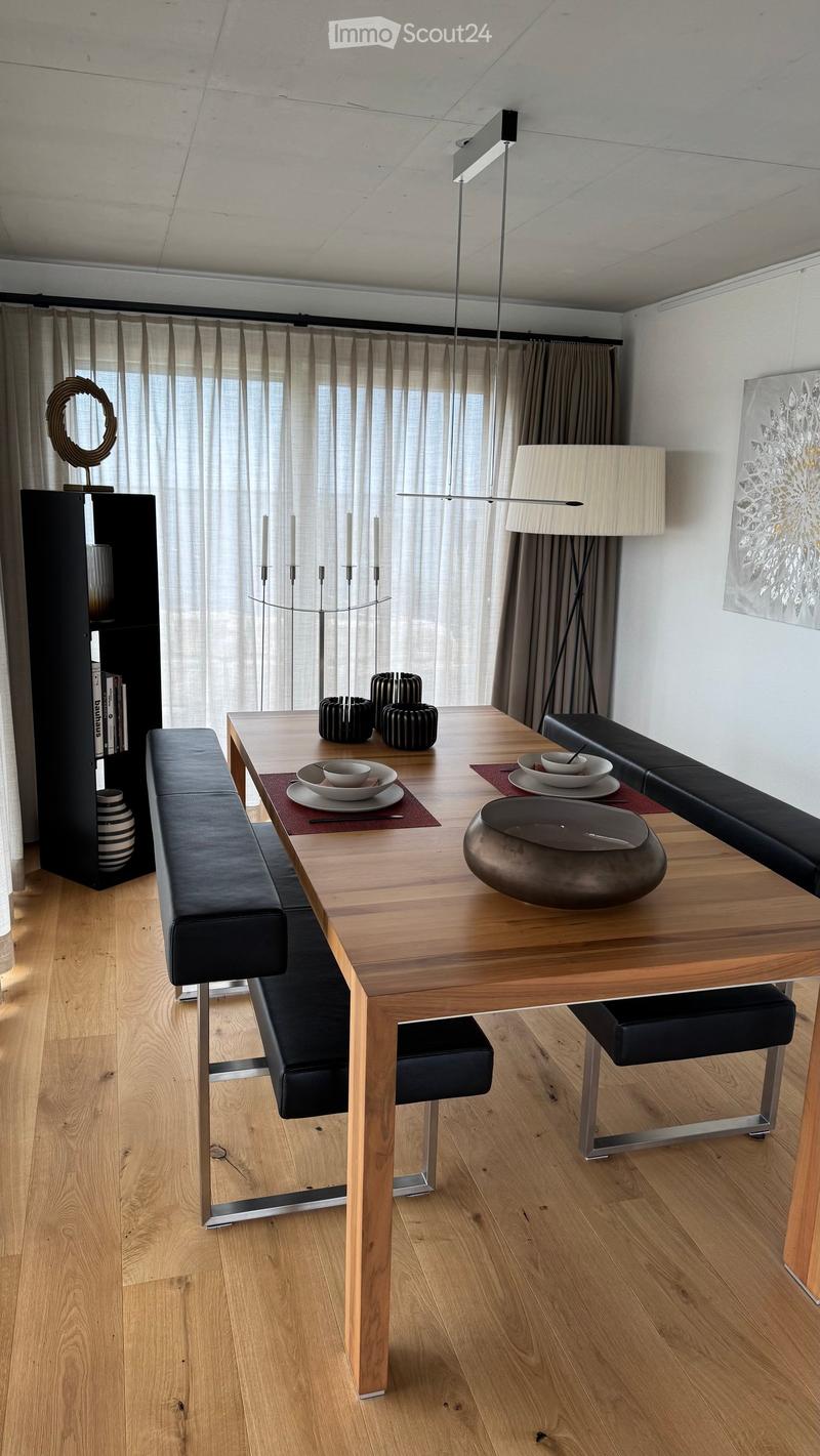 Modern dining area with a wooden table, black leather bench seating, decorative bowls, plates, and a sleek lamp. The room features large windows with sheer curtains and a contemporary aesthetic.