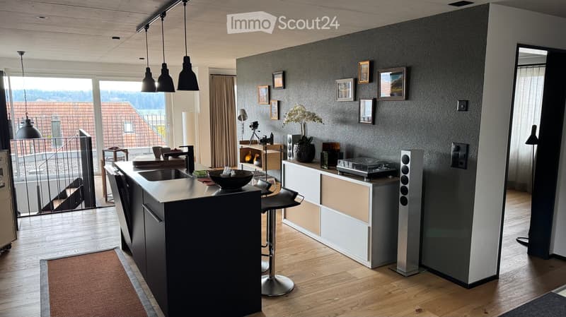 Modern kitchen with a central island, bar stools, and a sleek design. Features include a sink, pendant lights, a speaker system, and a view of the outdoor area.
