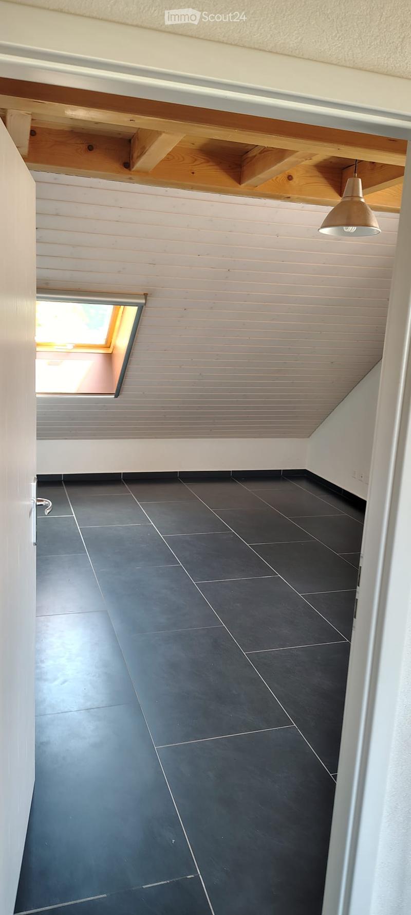 Empty room with dark grey tiled floor, white paneled walls, and a skylight window. Exposed wooden beams on the ceiling and a single pendant light fixture.