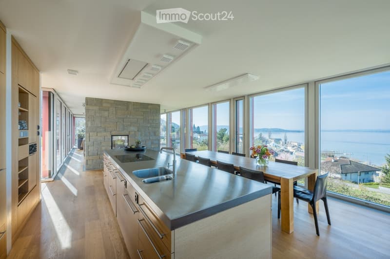 Modern kitchen with stainless steel island, sink, and faucet, wooden dining table with chairs, and a fireplace, overlooking a scenic view of water and landscape.