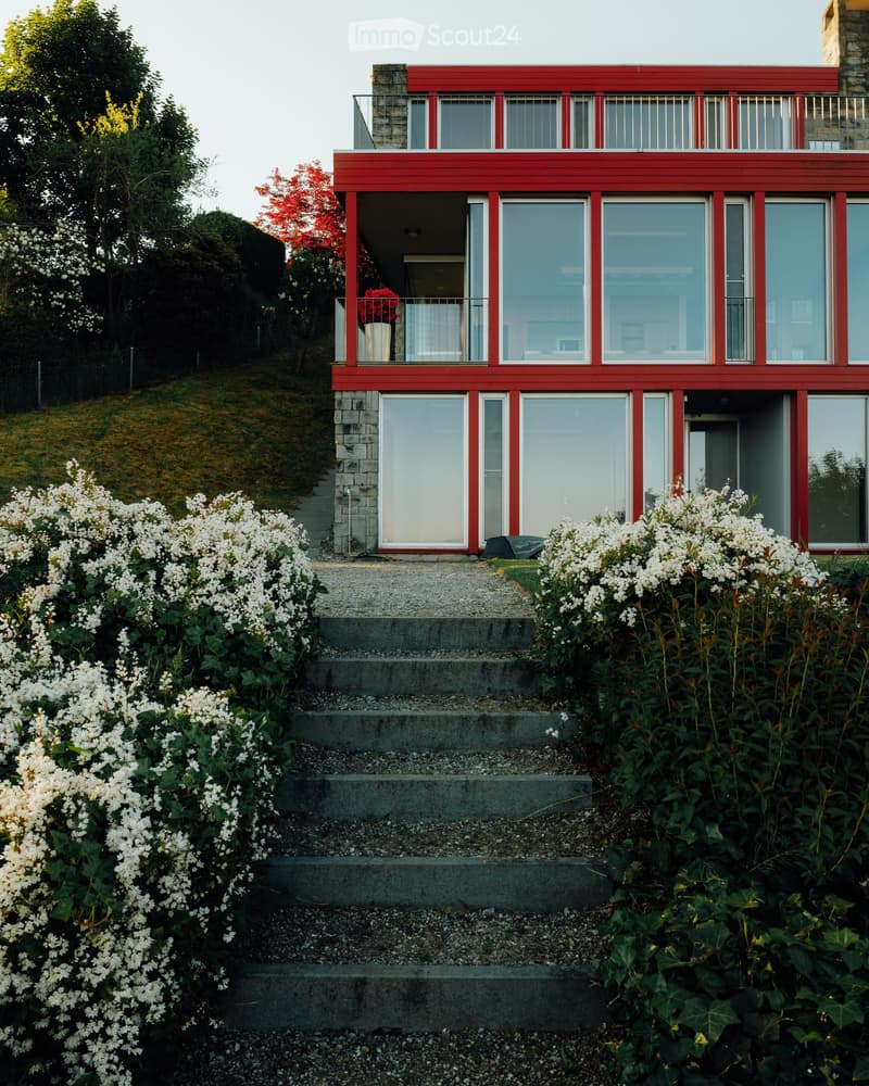 Two-story red house with large windows and a raised ground floor, surrounded by white flowering bushes and greenery.