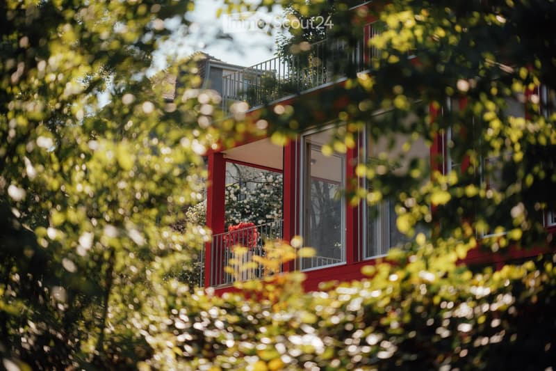 A modern red building with large windows and a balcony, partially obscured by lush green foliage and trees.