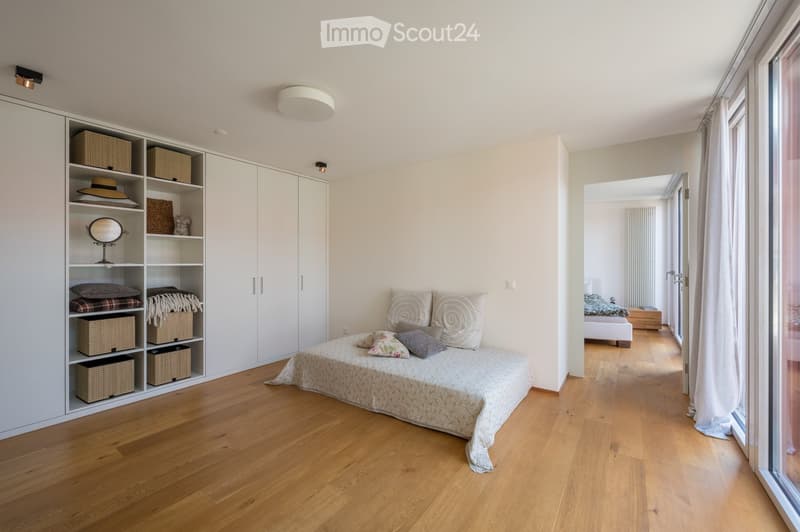 Modern bedroom with a large bed, decorative pillows, a built-in wardrobe with storage boxes, a mirror, and a hat on the shelf. The room features wooden flooring and large windows with curtains.