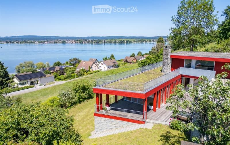 A modern red and white house with a green roof and balcony overlooking a lake, surrounded by trees and other houses.
