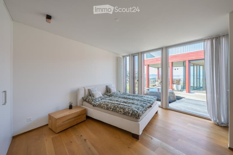 Modern bedroom with wooden flooring, large bed with patterned bedding, nightstand with lamp, large windows with curtains leading to a balcony area.