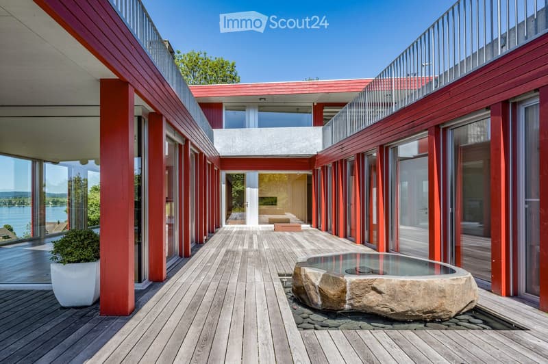 Modern outdoor deck area with wooden flooring, red wooden columns, glass railings, a stone water feature, and a view of a lake.