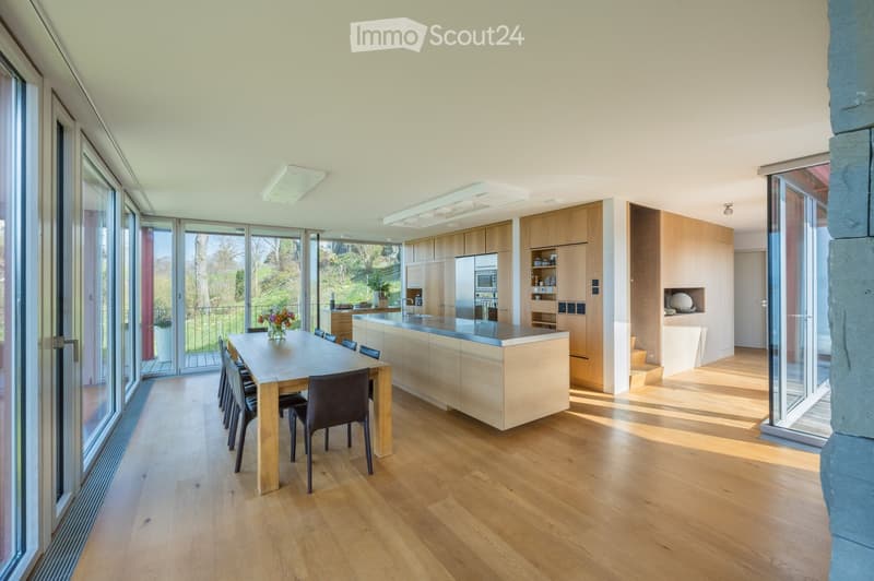Modern kitchen with light wooden cabinets, a central island, and a dining area with a wooden table and chairs. Large windows provide natural light and a view of the garden.
