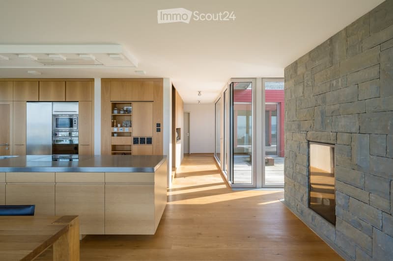 Modern kitchen with wooden cabinets, stainless steel appliances, a central island, and a stone fireplace. Large windows and sliding glass doors lead to an outdoor area.
