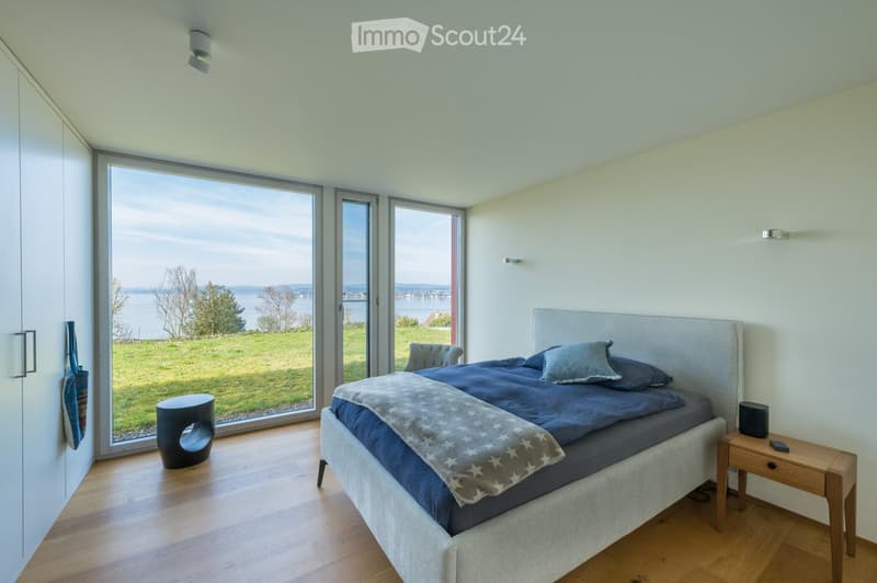 Modern bedroom with large windows offering a scenic view, featuring a bed with blue bedding, a nightstand, a stool, and a mirror.