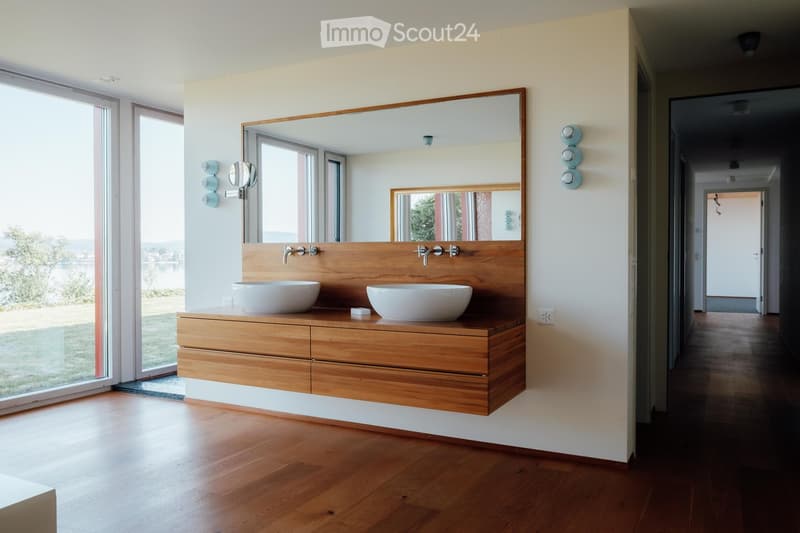Modern bathroom with wooden vanity, two white sinks, large mirror, and wall-mounted light fixtures. Large windows offer a scenic view.