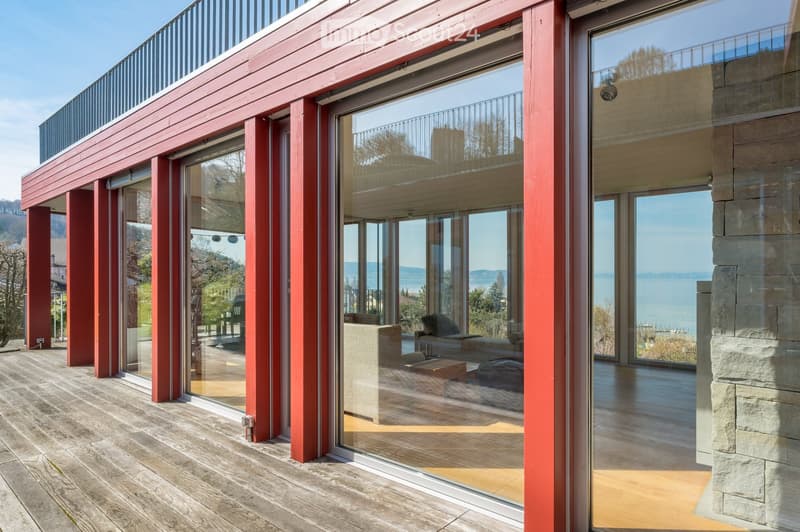 Modern balcony with large glass sliding doors, wooden decking, red wooden supports, and a view of the lake and surrounding landscape.