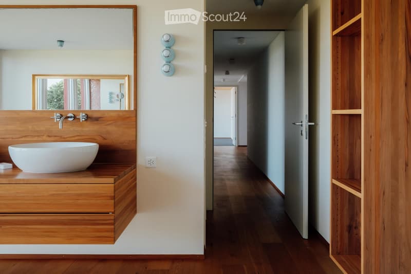 Modern hallway with wooden flooring, white walls, and recessed lighting. Features a wooden shelf on the right and a bathroom area visible on the left with a wooden vanity, sink, and mirror.