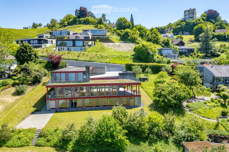 Aerial view of a modern, red-and-white house with large glass windows and a flat roof, situated on a hillside surrounded by lush greenery and other houses.