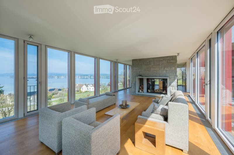 Modern living room with large windows offering a scenic view, a stone fireplace, a grey sofa, coffee table, armchairs, and an ottoman.