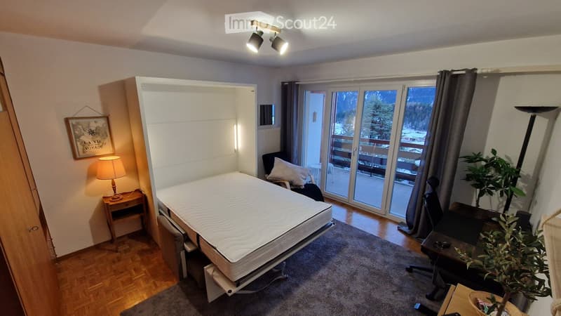 A room with a bed, a desk, a lamp, a picture frame, a potted plant, a chair, and a balcony with a view of the mountains.