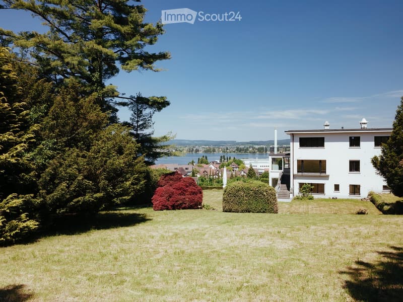 A modern white house with multiple windows and balconies, surrounded by a well-maintained lawn and trees, with a scenic lake view in the background.