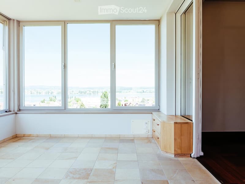 Bright, unfurnished room with large windows offering a scenic view, tiled floor, and a small wooden cabinet.
