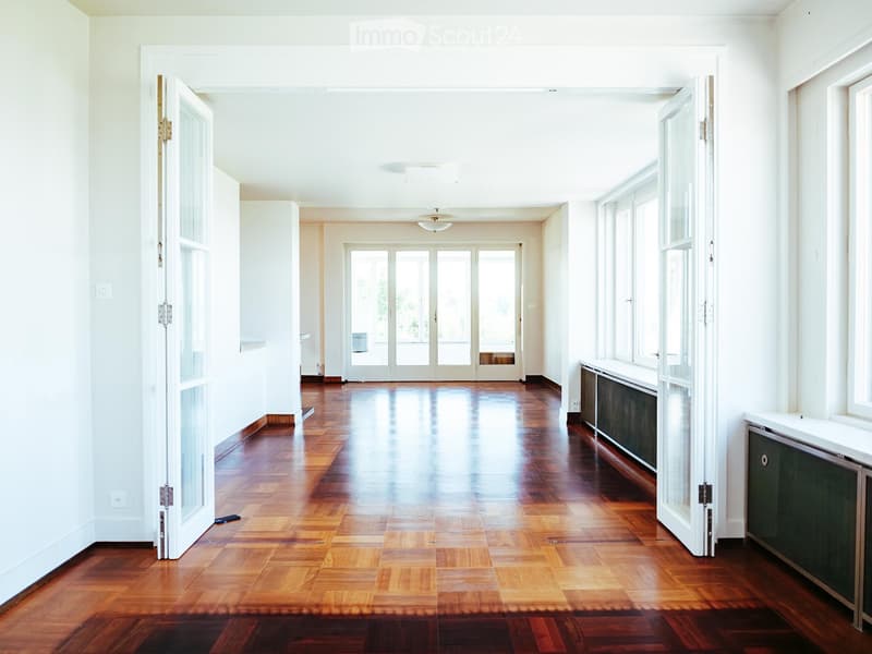 Spacious, well-lit hallway with polished wooden flooring, white walls, and large glass doors leading to another room.