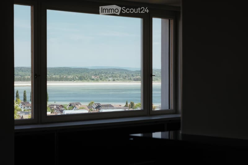 View from a large window showing a scenic landscape with a body of water, distant hills, and a town with houses.