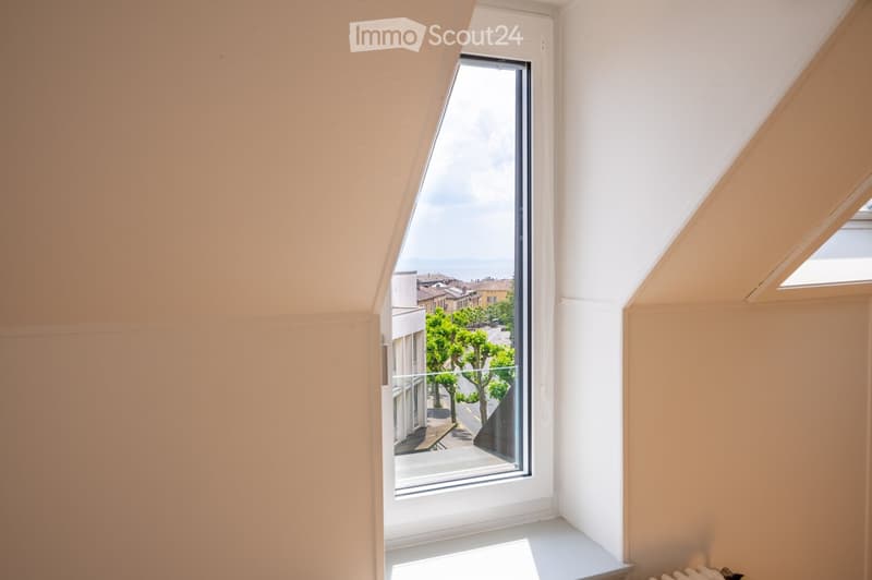 An empty room with light-colored walls and a large window offering a view of the street and trees. The room has a slanted ceiling typical of an attic space.