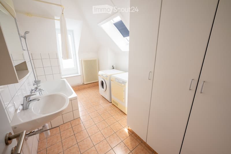Bright bathroom with a sink, bathtub, shower, radiator, and a private washing machine and dryer unit. The room features tiled flooring and a skylight window.