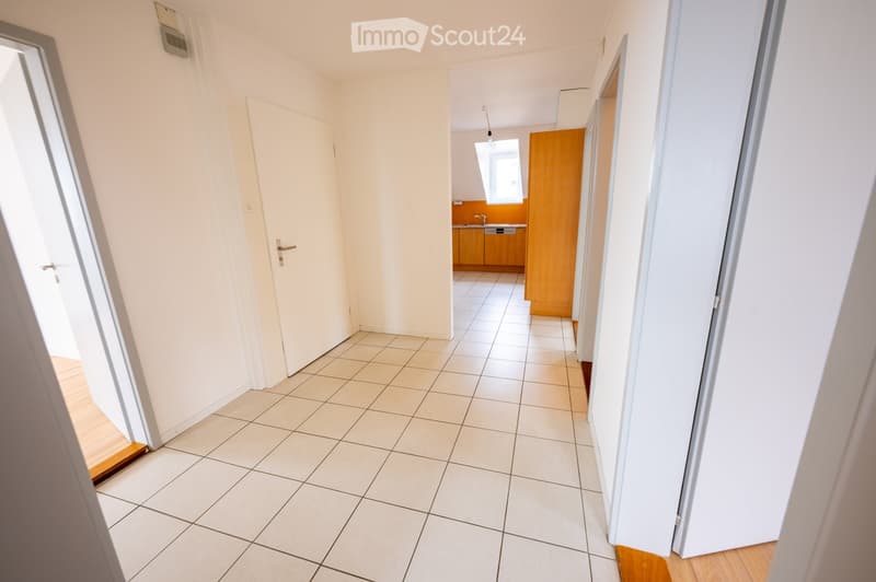 Empty hallway with tiled floor, white walls, and doors leading to other rooms, including a kitchen area with cabinets and a sink.