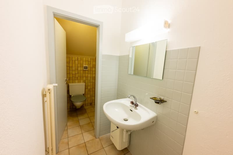 A small bathroom with a white tiled wall, a sink with a mirror above it, and a toilet in an adjacent room with tiled walls and floor.