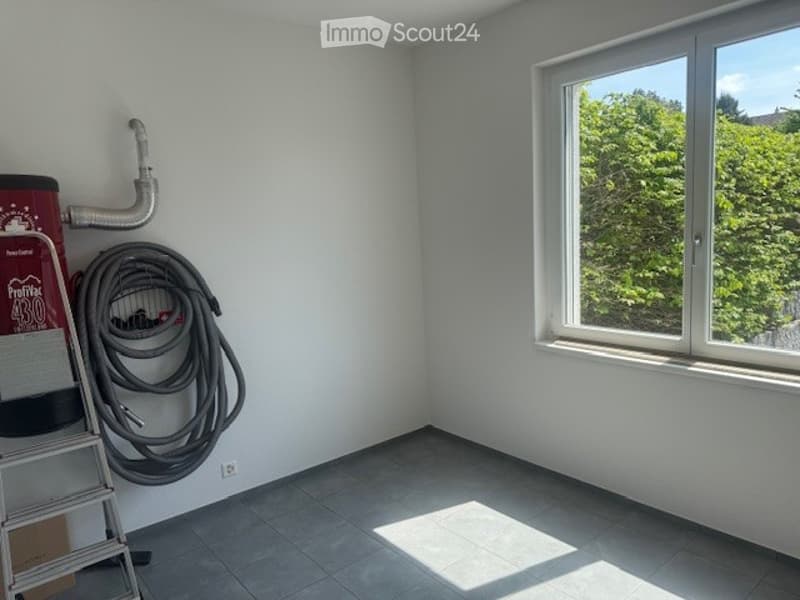 Empty room with white walls, tiled floor, and a large window showing greenery outside. A ladder and coiled hose are stored against the wall.