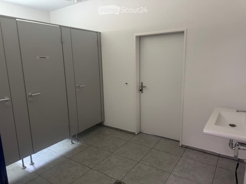 Empty restroom with tiled floor, white walls, two grey stall doors, a white sink, and a closed door.
