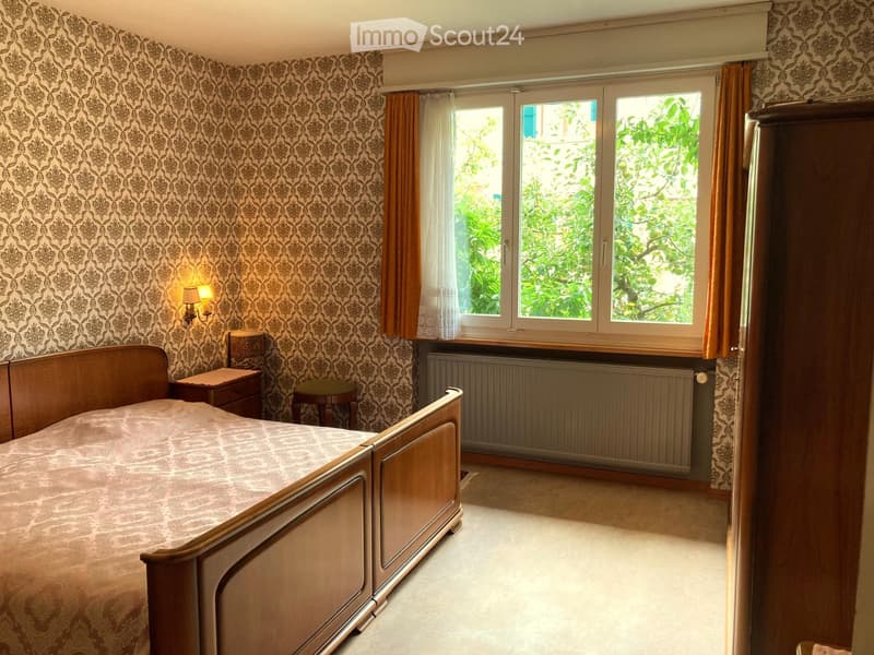 Bedroom with a double bed, nightstand, stool, lamp, and a wardrobe. The walls are covered with patterned wallpaper, and there is a window with curtains.