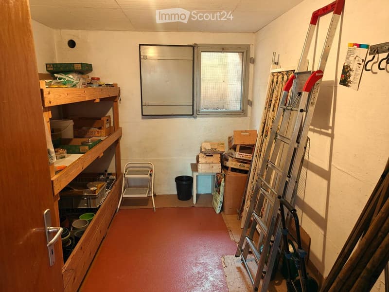 A storage room with wooden shelves, a ladder, a trash bin, and various storage boxes. The room has a red floor and a window with a metal grill.